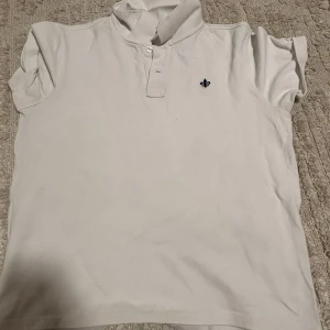 Vit pikétröja med broderad logga - Klassisk vit pikétröja med korta ärmar och krage. Tröjan har två knappar vid halsen och en liten svart broderad logga på bröstet. Tillverkad i mjuk bomull som känns skön mot huden. Perfekt för en clean och stilren look.