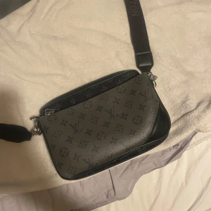 axelväska med monogram - Säljer en svart axelväska med klassiskt monogrammönster. Väskan har justerbar axelrem med logga, två fack med dragkedja och är tillverkad i skinn. Perfekt storlek för det viktigaste och riktigt snygg att bära crossbody.