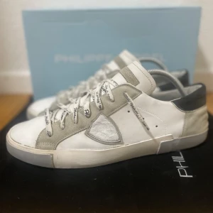 Philippe Model sneakers - Snygga vita sneakers från Philippe Model med detaljer i ljusgrå mocka och svart häl. Skorna har rund tå, platt sula och snörning med logotyp på skosnörena. Tillverkade i skinn och mocka för en exklusiv känsla. Bra skick! Sparsamt använda. Allt org medföljer. Nypris runt 3500 kr. Storlek 43. Hör av er ifall ni har några frågor! 💬 