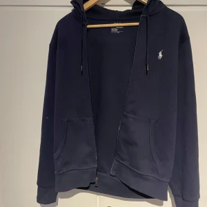 Mörkblå hoodie från Polo Ralph Lauren - Snygg mörkblå hoodie från Polo Ralph Lauren med dragkedja, huva och två stora fickor framtill. Klassisk broderad logga på bröstet och sköna muddar vid ärmslut och midja.