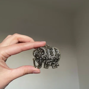 Vintage elefant-halsband  - Unikt halsband med stor elefantberlock i antik silverfärg. Berlocken är dekorerad med glittrande stenar och har ett detaljerat, snirkligt mönster. Perfekt statement piece för dig som gillar djurmotiv och bohemisk stil.