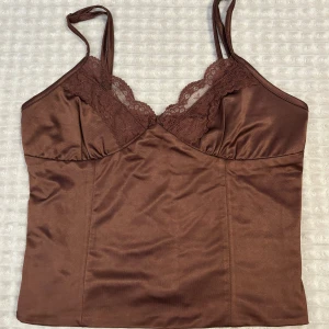 Brun satinlinnetopp från Bik Bok XS - Supersnygg brun linnetopp från Bik Bok i satin med tunna axelband och spetsdetaljer . Sitter figurnära och har en mjuk glansig finish. endast använd en gång för några år sedan. Inga skador. Storlek Xs