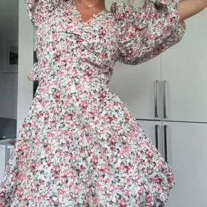 Blommig kortklänning med volang - Supersöt kortklänning med blommigt mönster i rosa, vitt och grönt. Klänningen har puffiga långärmar, v-ringning, volangdetaljer och snörning i midjan. Storlek M men passar bra på mig som har S då rosetten gör så den är justerbar. Nypris 399