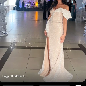 Champagnefärgad långklänning med slits - Superfin långklänning i champagnefärg med glittriga detaljer och offshoulder-ärmar. Klänningen har en djup slits framtill och en snygg omlottkänsla upptill. Perfekt för bal eller fest, ger en riktigt elegant vibe. Köpt på SE fashion i Södertälje använd en gång 