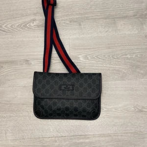 Gucci väska  - Snygg svart gucci väska med klassiskt GG-monogram över hela väskan. Väskan har en bred, justerbar rem i rött och marinblått med spänne. Perfekt storlek för det viktigaste och en stilren look.