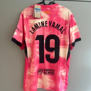 Barcelona Lamine Yamal rosa matchtröja - Säljer en Barcelona fotbollströja med Lamine Yamal och nummer 19. Tröjan är rosa och vit med tie-dye effekt, svarta detaljer och klubbmärke på bröstet. Nike-logga och Spotify som sponsor framtill. Tillverkad i lätt och ventilerande material.