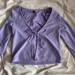 Ljuslila ribbad omlottopp med knyt - Superfin ljuslila omlottopp med ribbad struktur och långa ärmar. Toppen har en snygg v-ringning och knyt framtill som ger en trendig look. Croppad modell som passar perfekt till höga jeans eller kjol.