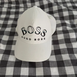 Vit keps från Hugo Boss - Snygg vit keps från Hugo Boss med broderad BOSS-logga framtill i svart och turkos. Justerbar rem baktill med metallspänne där det står BOSS. Klassisk böjd skärm och ventilerande hål på sidorna. Perfekt accessoar för en clean och stilren look.