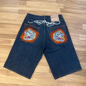 Ed Hardy jorts med tigerbrodyr - 🚨Pris kan diskuteras 🚨Unika mörkblå jeansshorts från Ed Hardy med stora broderade tigrar i orange, blått och vitt på bakfickorna. Snygg Ed Hardy-logga broderad ovanför fickorna. Klassisk femficksmodell i denim med loose fit och orange kontrastsömmar.                     