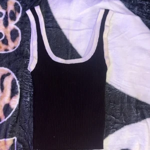 Svart ribbad linnetopp med vit kant - Snygg svart ribbad linnetopp med breda axelband och kontrasterande vit kant runt halsringning och ärmhål. Perfekt att styla med jeans eller kjol för en cool och enkel look.