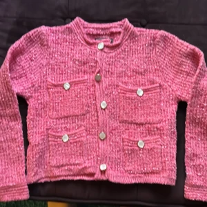 Rosa Lunetta  jacka från Alessa - Superfin rosa cardigan jacka från Alessa med glittriga inslag och snygga pärlemorknappar. Jackan har fyra fickor framtill och är stickad i ett mjukt material. Perfekt för dig som vill sticka ut med en färgglad och trendig look.