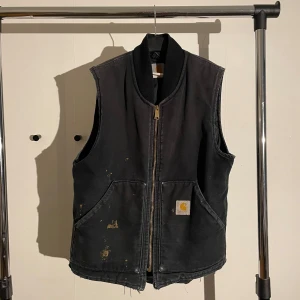 Vintage(90s) Svart Carhartt dunväst med dragkedja - Svart dunväst från Carhartt med grov guldfärgad dragkedja, två stora fickor framtill och ribbad krage. Västen har quiltat foder och klassisk Carhartt-logga på ena fickan. Västen passar S/M. Västen är från 90-tal och nyligen tvättad. Vid fler frågor eller bilder, tveka ej att skriva! 