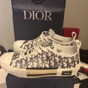 Dior B23 sneakers med monogram - Snygga Dior B23 sneakers med ikoniskt grått och vitt monogrammönster, vita snören och detaljerad sula med Dior-logga. Skorna har genomskinliga paneler och klassisk rund tå. Perfekt för dig som vill sticka ut med lyxig streetstyle. Tveka inte med o skriva.