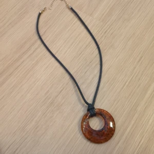 Coolt halsband med berlock - Unikt halsband med svart snöre och en rund, brunmelerad hängberlock. Hängberlocken har en retro vibe och gör alla outfits snygga. Säljer eftersom det tyvärr inte kommer till användning. 