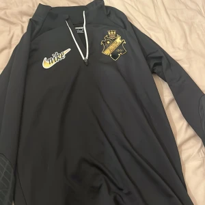 AIK x Nike svart Dri-FIT half zip tröja - Svart långärmad AIK-tröja från Nike med guldiga detaljer och klubbmärke på bröstet. Slim fit-modell i Dri-FIT-material som andas, perfekt för fotbollsträning. Halv dragkedja framtill och snygg vit kant vid zippen. Grym för dig som vill representera AIK på planen. Helt oanvänd och perfekt skick. Riktigt fet AIK zip från AIK shop i storlek S. 