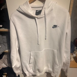 Vit hoodie från Nike med logga - Snygg vit hoodie från Nike med klassisk svart logga på bröstet. Tröjan har huva med snörning, magficka och långa ärmar. Perfekt för chill dagar eller när du vill ha en clean och enkel stil. Mjuk och skön i bomullsmix.