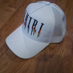 Vit meshkeps från Amiri - Snygg vit keps från Amiri med broderad logga framtill i svart och färgglada detaljer. Kepsen har meshpaneler på sidorna och bak för extra ventilation samt justerbar snapback. Perfekt för en streetwear-look.