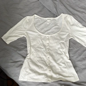 Vit ribbad topp med knappar - Superfin vit topp med ribbat tyg och smala vertikala ränder. Modellen har trekvartsärm, djup rundringning och knappar framtill. Perfekt att styla med jeans eller kjol för en clean look.