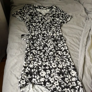 Svartvit blommig omlottklänning Ellos - Svartvit kortklänning från Ellos med blommigt mönster, v-ringning och knytband i midjan. Klänningen har korta ärmar och är tillverkad i ett mjukt, lätt material som passar perfekt för varmare dagar.