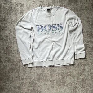 Vit sweatshirt från Hugo Boss - Vit sweatshirt från Hugo Boss med broderad logga i blått och turkost framtill. Tröjan har rund hals, långa ärmar och ribbade muddar. Skön och stilren modell i mjuk bomull, perfekt för en clean och avslappnad look.