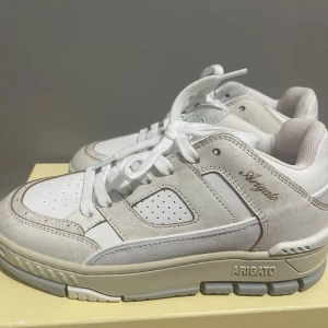 Vita sneakers Axel Arigato - Snygga vita sneakers från Axel Arigato med chunky sula och detaljer i mocka och skinn. Skorna har perforerade partier, vita snören och diskret Arigato-logo i guld på sidan. Perfekt för dig som gillar stilrena och trendiga sneakers.