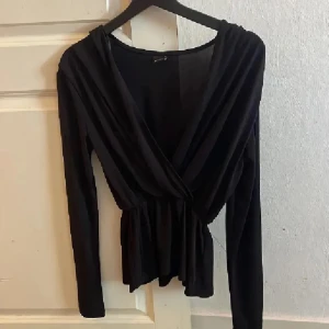 Svart omlottopp från Gina Tricot - Svart omlottopp från Gina Tricot i mjuk polyester. Toppen har lång ärm, v-ringad omlottfront och smickrande rynk vid midjan. Perfekt att styla med jeans eller kjol för en cool look. Snyggt flowy tyg och enkel att matcha med accessoarer.