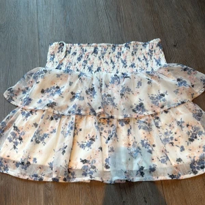 Blommig volangkjol från Vero Moda - Supersöt kort kjol från Vero Moda med blommigt mönster i blått och rosa på vit botten. Kjolen har bred smock i midjan och två lager volanger som ger en flowy och romantisk vibe. Perfekt för dig som gillar en feminin och fräsch stil.