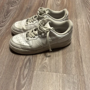Nike Air Force 1 vita sneakers - Säljer ett par klassiska Nike Air Force 1 sneakers i helvitt skinn. Modellen har perforerad tå, snörning och platt sula. Perfekt för dig som gillar tidlös streetstil och vill ha ett par sneakers som funkar till allt. Ikonisk siluett och bekväm passform.