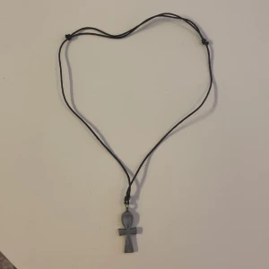 Svart halsband med ankh-symbol - Coolt svart halsband med en grå ankh-symbol som hänge. Halsbandet har justerbar snodd.