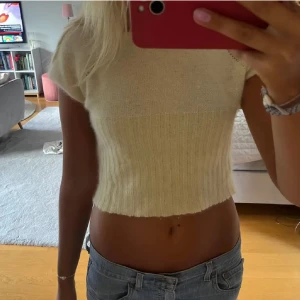 Beige croppad stickad topp - Superfin croppad topp i beige stickat material med korta ärmar och ribbad nederdel. Den har en enkel, clean look och är perfekt att matcha med jeans eller kjol. Mjuk och skön, med en lätt oversized känsla över axlarna.