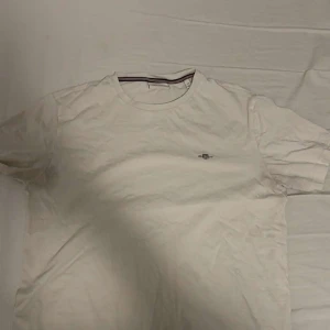 Vit t-shirt från Gant - Snygg vit t-shirt från Gant i klassisk modell med rund hals och diskret logga broderad på bröstet. Halsen har en cool detalj med röd, vit och blå rand på insidan. Perfekt för en clean och stilren look.