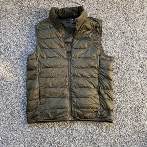 Olivgrön dunväst från Uniqlo - Säljer en olivgrön dunväst från Uniqlo Ultra Light Down. Västen har quiltad design, hög krage och dragkedja framtill. Den är superlätt och har en clean, sportig look som funkar till många outfits. Perfekt för lager-på-lager under kyliga dagar.