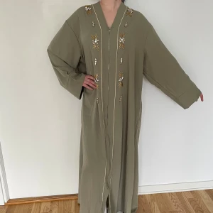 Olivgrön långklänning med pärlor - Snygg olivgrön långklänning med breda ärmar och dekorativa pärlor och broderier i guld och silver längs framsidan. Klänningen har en rak, loose passform och dragkedja framtill. Perfekt för dig som gillar unika detaljer och en chill vibe. Abaya
