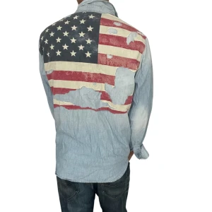 Ralph Lauren denim supply skjorta  - Säljer en ljusblå jeansskjorta från Ralph Lauren med lång ärm och knappar framtill. På ryggen finns en stor amerikansk flagga i rött, vitt och blått med slitna detaljer. Skjortan har två bröstfickor med lock och en liten logotyp på vänster sida.