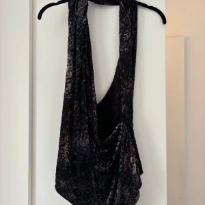 Svart omlott halterneck topp i sammet - Säljer en svart omlott halterneck topp i mjuk sammet med snygg drapering framtill. Toppen är ärmlös och har öppen rygg som ger en cool och trendig vibe. Perfekt för dig som gillar att sticka ut med en unik och lyxig look.