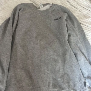 Grå sweatshirt från Bula i bomull - En enkel och stilren grå sweatshirt från Bula i storlek M. Tröjan har rund hals, långa ärmar och ett litet Bula-tryck på bröstet. Mjuk insida och ribbade muddar vid hals, ärmslut och nederkant. Perfekt för chill dagar eller när du vill ha något bekvämt.