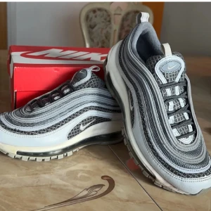 Nike Air Max 97 svart och grå sneakers - Nike Air Max 97 sneakers med vågiga linjer i svart och grått, och coolt reptilmönster. Skorna har synlig Air-sula, snörning och rund tå. Köpta på JD och finns inte längre i produktion. Perfekt för dig som gillar streetstyle och sportig vibe.