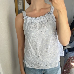 Ljusblå broderad topp med volang - Supersöt ljusblå topp med breda axelband och volangkant upptill. Toppen har broderade bladmönster och små hålbroderier som ger en luftig känsla. Perfekt för varma dagar och enkel att styla med jeans eller shorts. Storlek 13-14 barn men passar xs/s!