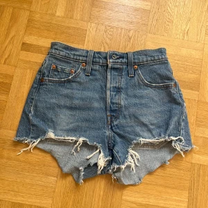 Levi's blå jeansshorts med fransar - Säljer ett par klassiska blå jeansshorts från Levi's med hög midja och råa, fransiga kanter. Modellen har fem fickor, bälteshällor och Levi's ikoniska läderpatch bak. Perfekt för dig som gillar en avslappnad och edgy stil. Aldirg använda. Skriv vid minsta fråga!💕💕