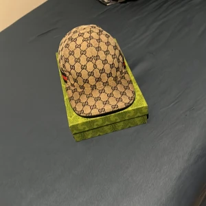 Gucci keps med GG-mönster och röd-blå rand - Snygg keps från Gucci i beige med klassiskt GG-monogram över hela kepsen. Har en röd och blå rand på sidan och justerbart spänne bak. Materialet är canvas med insida i mörk textil. Perfekt för dig som vill sticka ut med en lyxig accessoar.