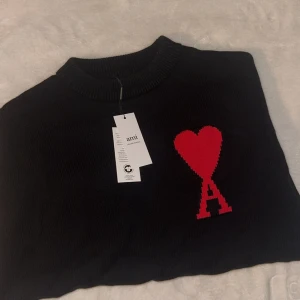Svart stickad tröja från Ami med hjärta - Svart stickad tröja från Ami med ett stort rött hjärta och bokstaven A på bröstet. Tröjan har ribbad rund hals och muddar vid ärmslut och nederkant. Perfekt för dig som gillar stilrena plagg med en cool detalj. Passar dig som vill ha något unikt i garderoben. Tröjan är storlek M och säljer den för den passar ej mig