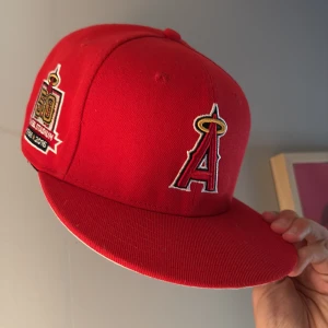 Röd LA Angels keps från New Era - Snygg röd 59FIFTY keps från New Era med LA Angels logga framtill och broderad patch på sidan för 50-årsjubileum. MLB-logga baktill och platt skärm. Tillverkad i 100% polyester, perfekt för dig som gillar baseball och streetwear.