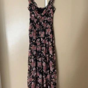 Svart blommig långklänning med volang - Supersöt långklänning med svart botten och rosa blommigt mönster. Klänningen har volangdetaljer på axelbanden och smock över byst och midja för en skön passform. Materialet är lätt och luftigt, perfekt för varma dagar. Axelbanden är breda och har fina ryschkanter.