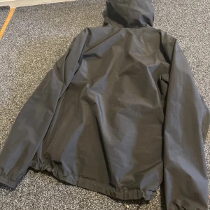 Svart Haglöfs Gore-Tex vindjacka - Svart vindjacka från Haglöfs med Gore-Tex-material, perfekt för blåsiga och regniga dagar. Jackan har huva, två dragkedjeförsedda fickor och meshfoder på insidan. Snygg minimalistisk design med logga på bröstet och elastisk nederkant.