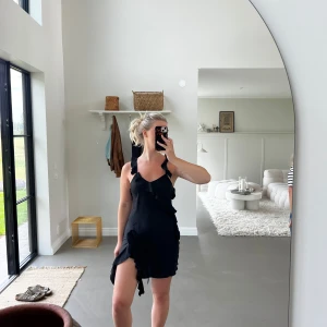 Svart omlottklänning med volanger - Säljer en svart kort omlottklänning med volangdetaljer och smala axelband som korsas i ryggen. Klänningen har en lätt asymmetrisk nederkant och öppen rygg, vilket ger en cool och feminin vibe. Perfekt för dig som gillar stilrena och trendiga plagg.