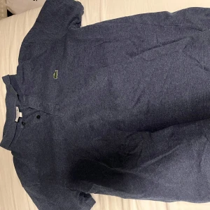 Blå Lacoste slim fit pikétröja - Snygg blå pikétröja från Lacoste i slim fit-modell. Klassisk krage, två knappar och den ikoniska gröna krokodilloggan på bröstet. Materialet är mjuk bomull som känns skönt mot huden. Perfekt för dig som gillar stilren och sportig vibe.