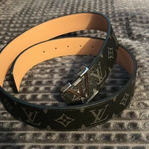 Louis Vuitton svart skinnbälte - Snyggt svart bälte från Louis Vuitton med klassiskt monogrammönster och en stor LV-spänne i silver. Bältet är gjort i skinn med beige insida och har en stilren, lyxig känsla som verkligen sticker ut. Perfekt för dig som vill ha något extra till outfiten.