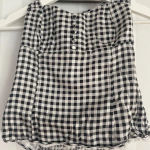 Svartvit rutig linnetopp med knyt - Snygg linnetopp från H&M i svartvitt rutigt mönster. Toppen har knyt i nacken, smockad rygg och dekorativa knappar framtill. Materialet är mjukt och lätt, perfekt för varma dagar. Passformen är normal och toppen är ärmlös.