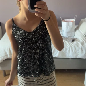 Svart glittrig linnetopp med paljetter - Svart linnetopp med tunna axelband och massor av glittrande paljetter som ger en riktigt festlig vibe. Toppen har v-ringad hals och ärmlös design, perfekt för att sticka ut på kvällen. Snygg att matcha med jeans eller kjol för en cool look.