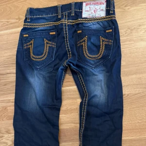 True Religion jeans straight baggy - Snygga blå jeans från True Religion med ikoniska kontrastsömmar i gult och stora broderade bakfickor. Klassisk femficksmodell med silverfärgade knappar och patch bak i midjan. Jeansen har en rak passform och cool tvättad look.